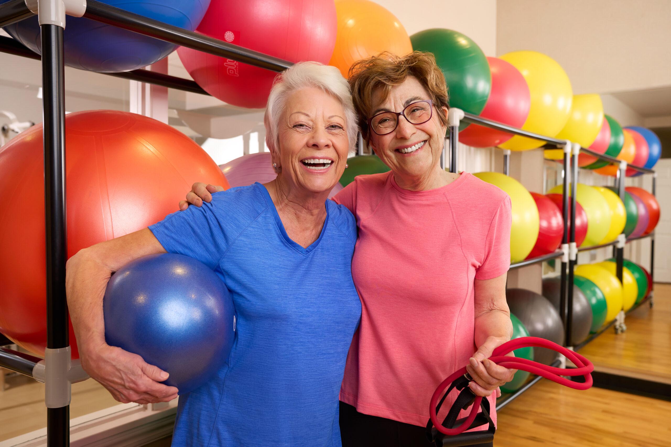 Two older woman smile with workout equipment in front of colorful exercise balls