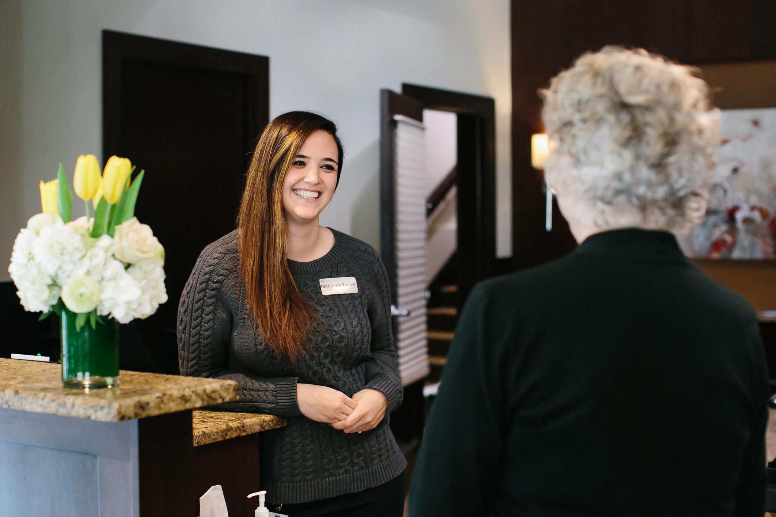 A smiling front desk clerk talks with an older person who is facing away from the camera