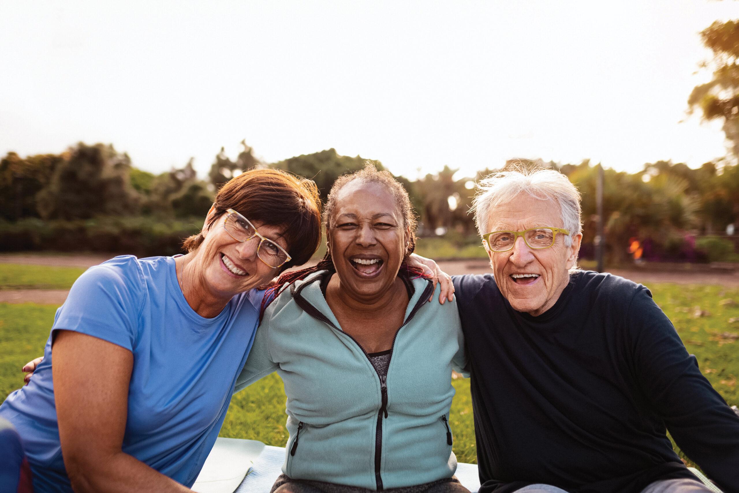 A group of three friends smiles outdoors while sitting on a picnic blanket or mat.