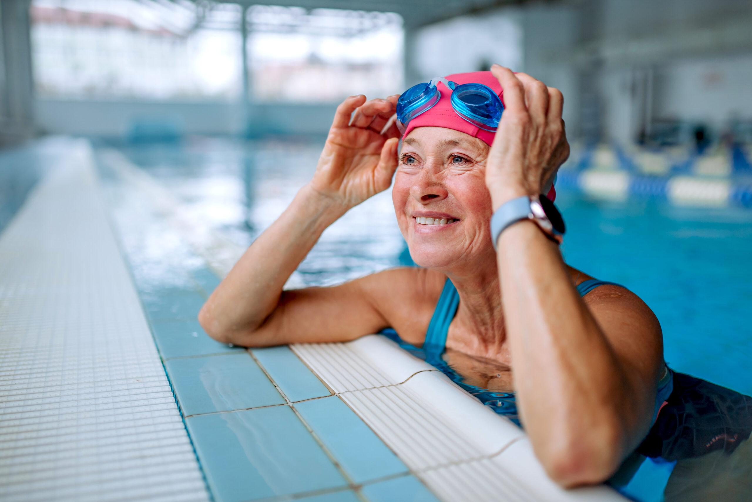 A smiling woman leans on the edge of the pool as she adjusts her goggles.