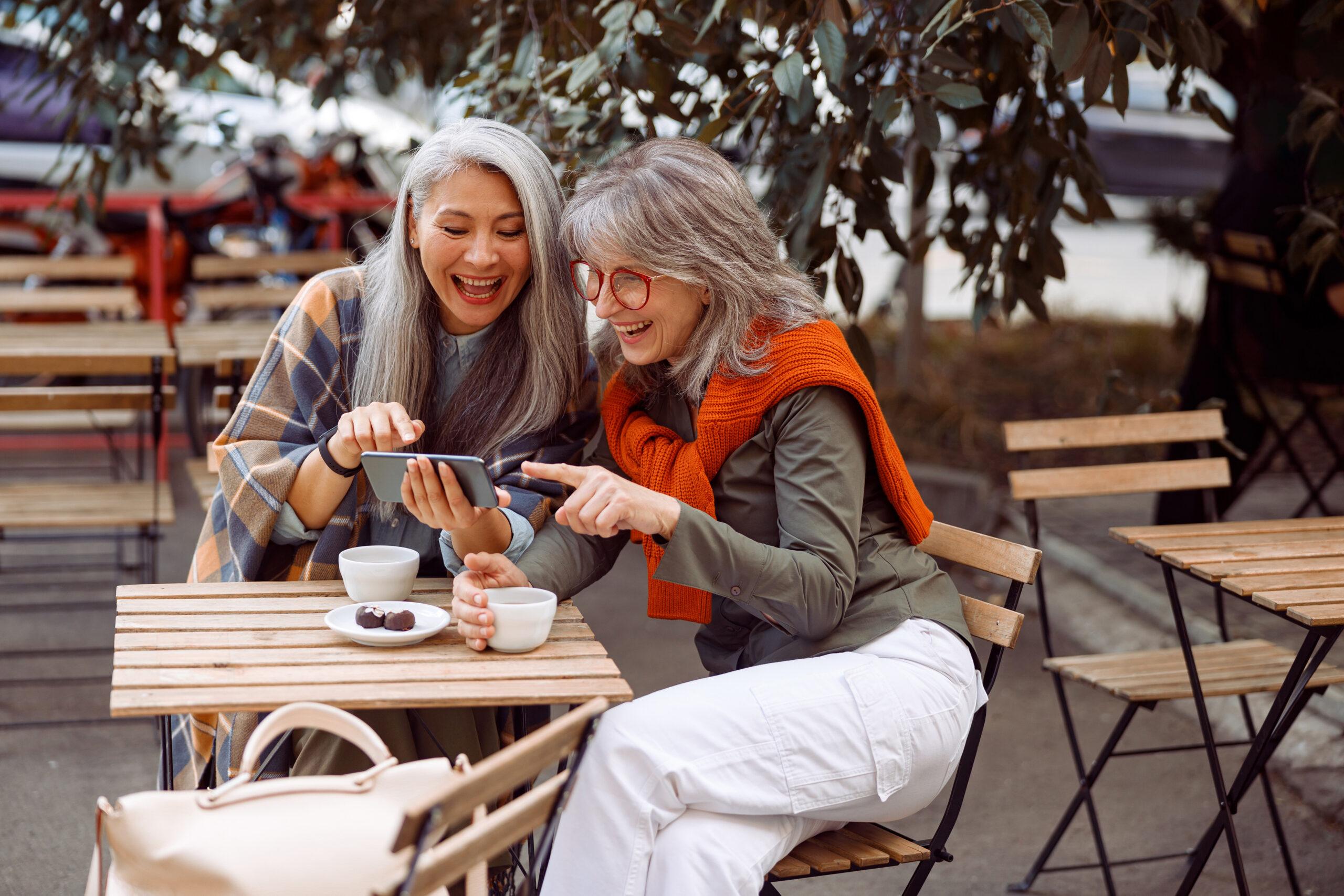 Two older women smile and look at a smartphone together at a cafe