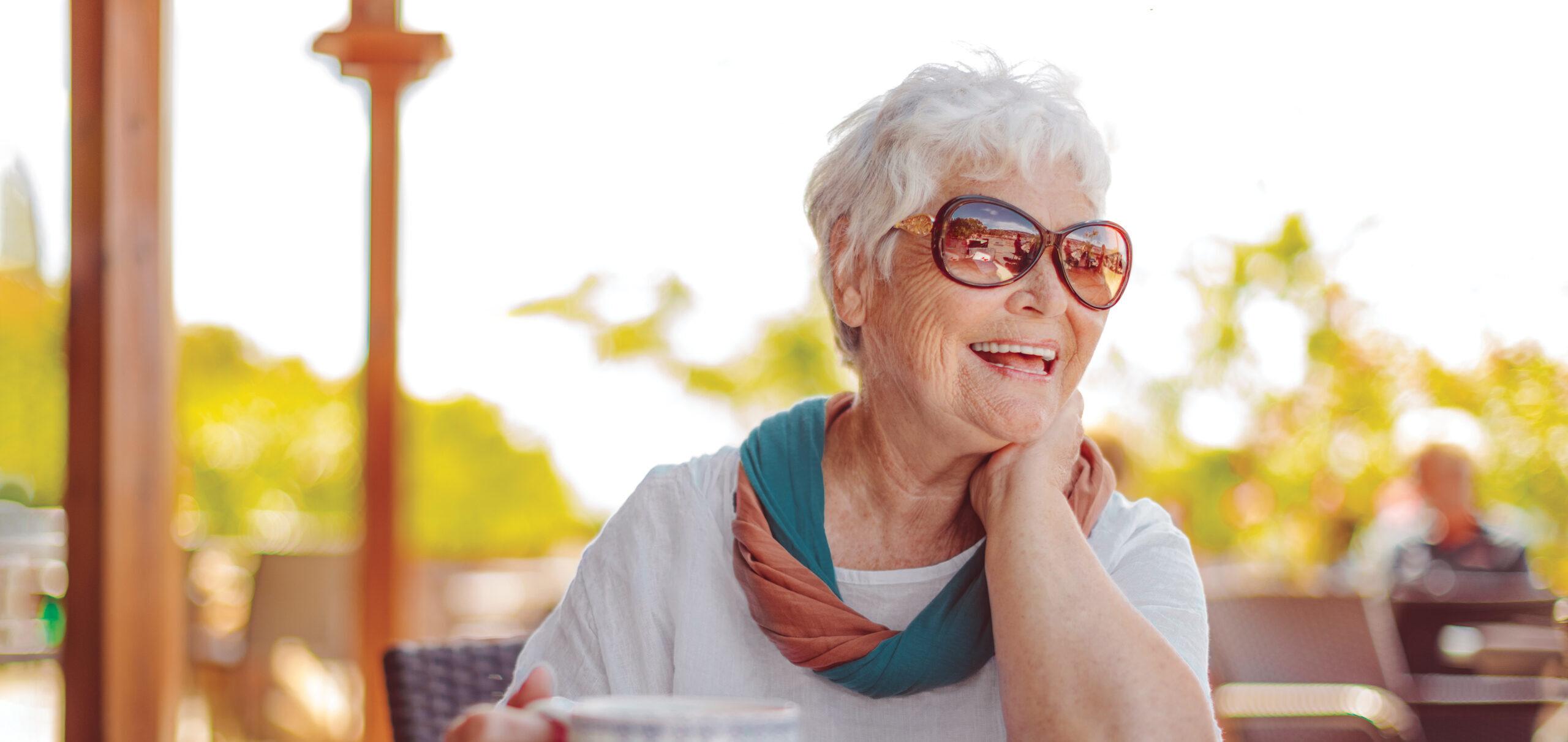 An older woman wearing sunglasses smiles with a hot beverage outdoors