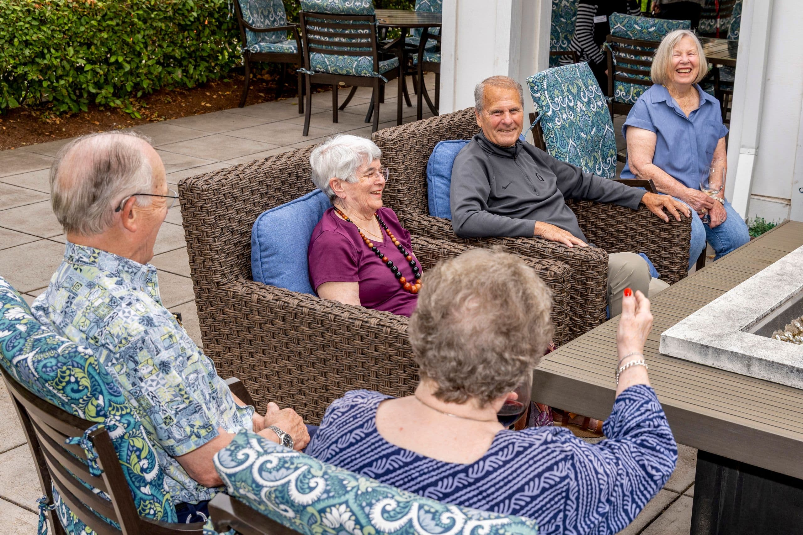 Five older adults are seated on an outdoor patio, laughing and talking around a table with a fire pit.