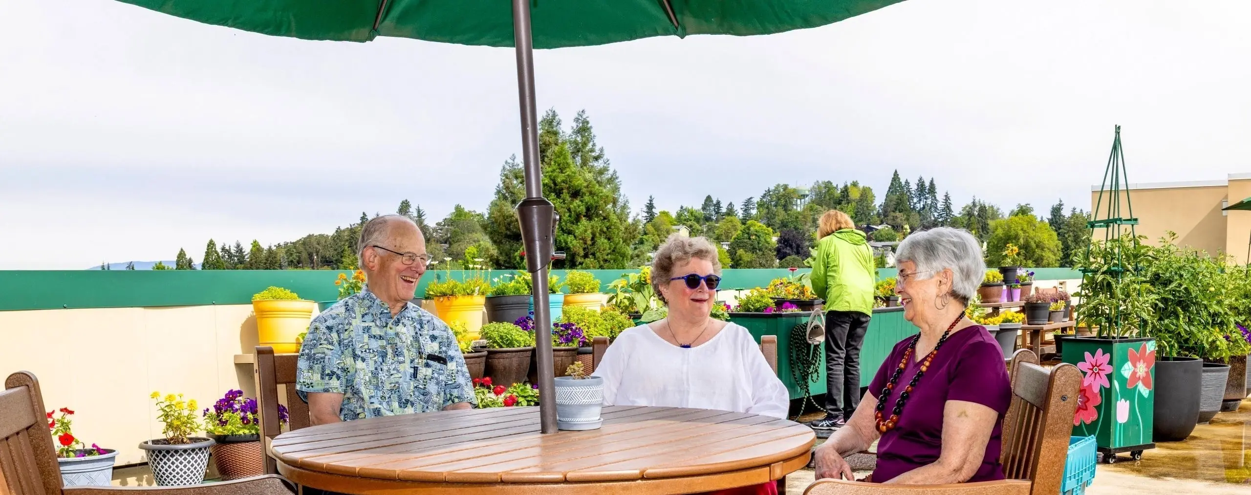Three smiling older adults sit at a table on a rooftop garden, surrounded by potted plants. Another person tends to plants in the background.