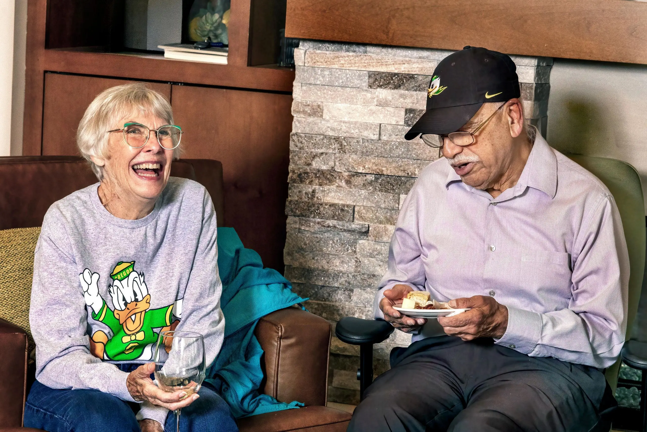 An woman laughing while holding a wine glass, and a man looking at a plate of snacks, both seated indoors.
