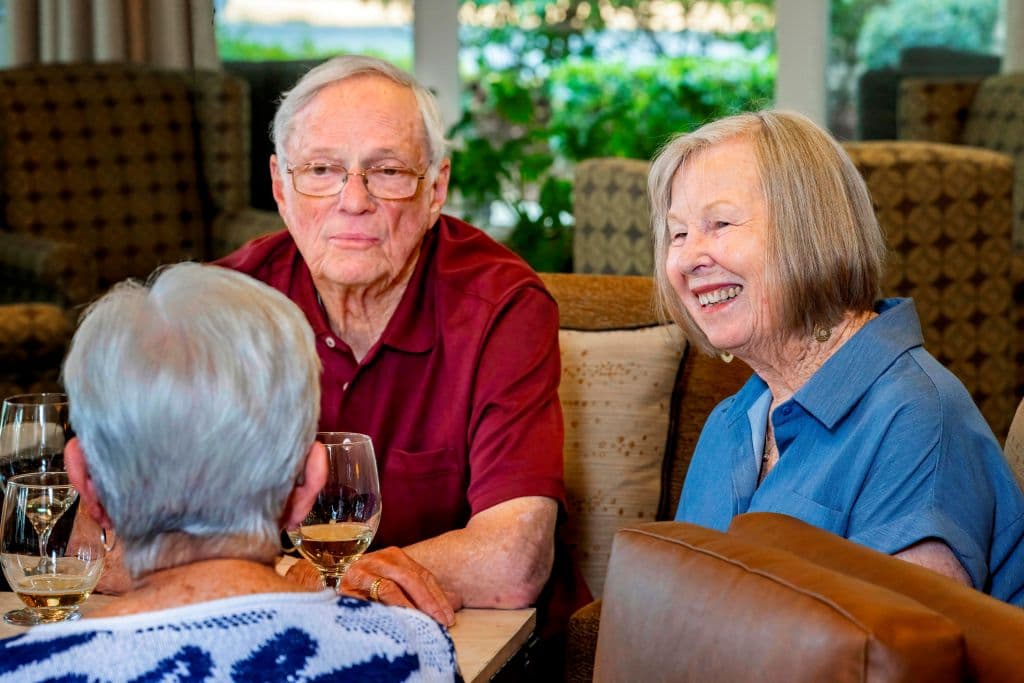 Three people sitting around a table with drinks. One woman smiles broadly, another man looks forward, and a third person has their back to the camera.
